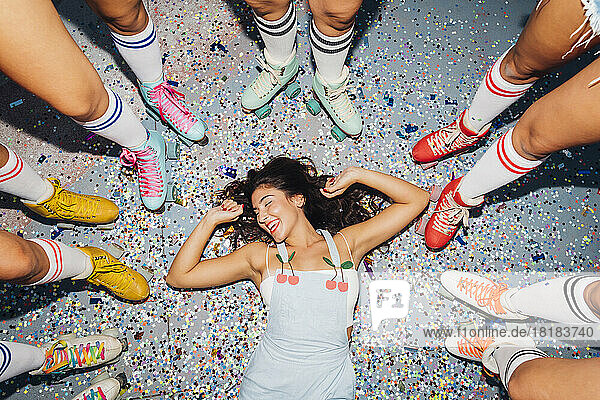Happy young woman lying on confetti amidst legs of of friends wearing roller skates