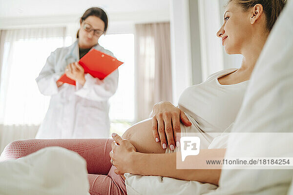 Smiling pregnant woman lying on bed with doctor in background