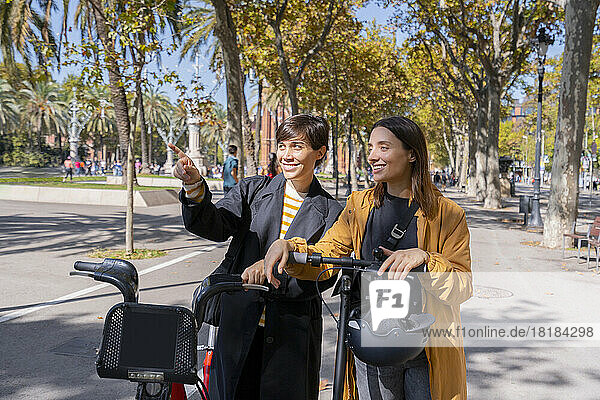 Lächelnde Freunde mit Elektrofahrrad und Tretroller diskutieren auf Fußweg