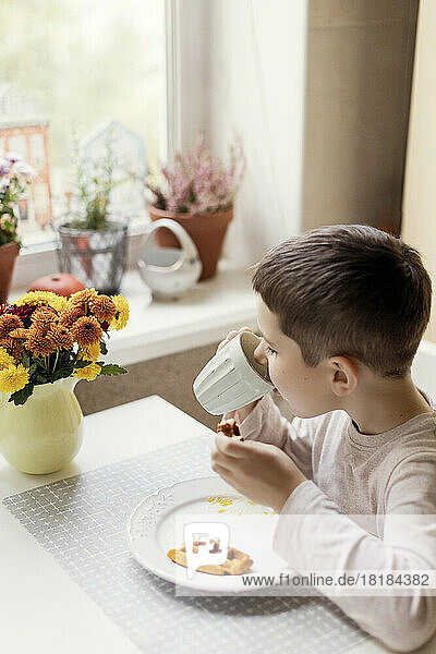 Boy drinking hot chocolate from cup on dining table in kitchen