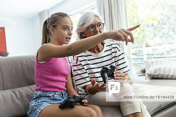 Granddaughter pointing by grandmother sitting with joystick on sofa at home