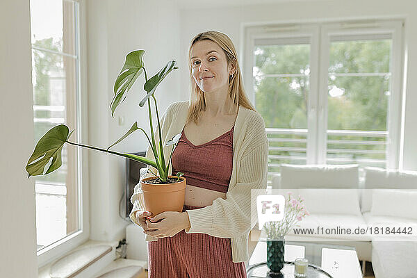 Smiling young woman with monstera plant standing in living room at home