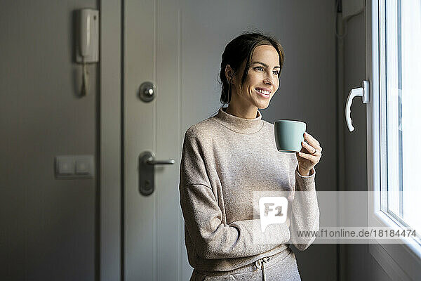 Happy woman holding coffee cup standing near window at home