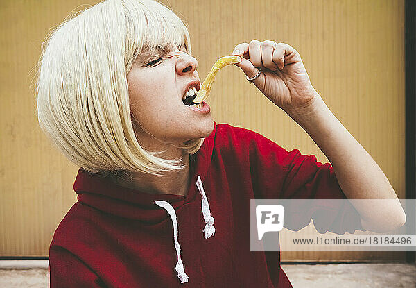 Blond woman eating French fries in front of brown wall