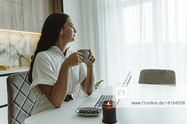 Businesswoman holding coffee cup sitting with laptop on table at home