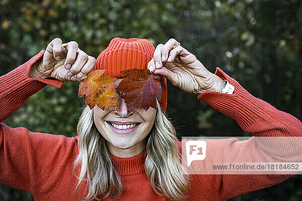Lächelnde Frau bedeckt am Wochenende die Augen mit Herbstblättern