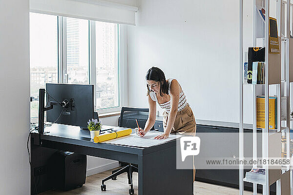 Young architect leaning on desk analyzing blueprint in office