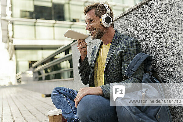 Smiling man talking on speaker phone sitting by wall