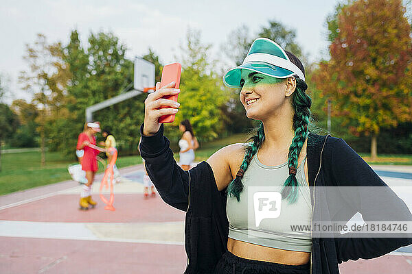 Lächelnde Frau mit Sonnenblende macht Selfie mit dem Handy auf dem Sportplatz