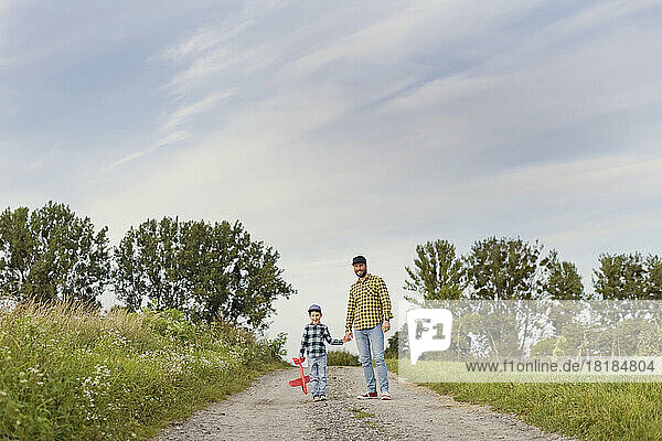 Boy with toy airplane holding hand of father on dirt road