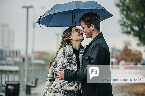 Smiling young man and woman kissing under umbrella on footpath