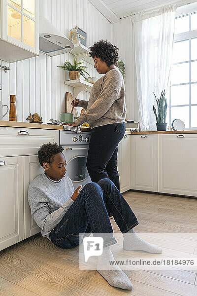 Mutter bereitet Essen zu  während ihr Sohn zu Hause auf dem Boden neben dem Schrank in der Küche sitzt