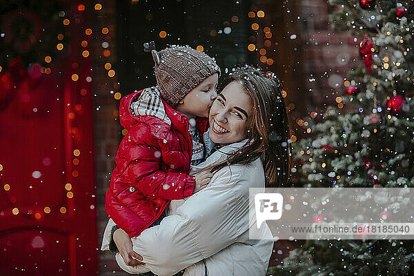Boy kissing mother standing in front of Christmas tree in snow