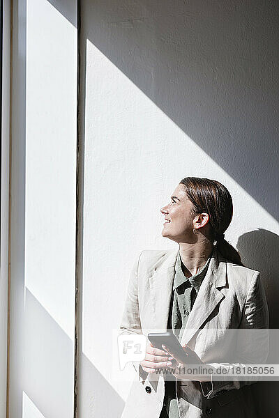 Happy businesswoman holding mobile phone leaning on wall