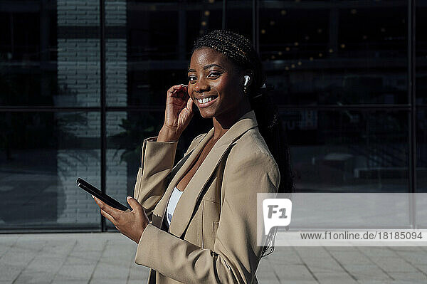 Lächelnde Geschäftsfrau mit Smartphone und kabellosen In-Ear-Kopfhörern am Fußweg