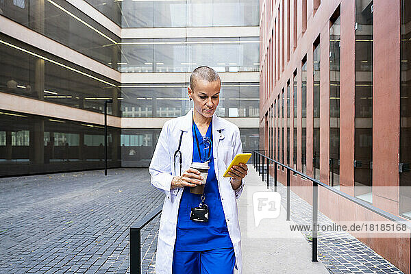 Mature doctor with disposable coffee cup using smart phone on footpath
