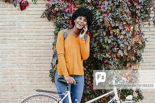 Happy woman with bicycle talking on smart phone in front of ivy wall