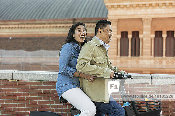 Happy mature couple riding electric bicycle by wall