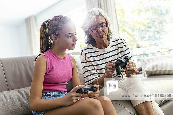 Grandmother showing game controller to granddaughter sitting on sofa at home