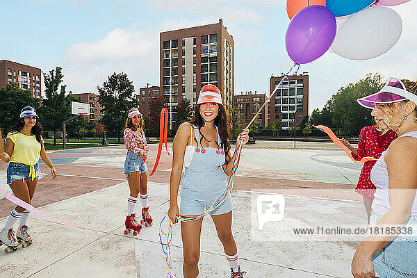 Smiling woman holding balloons standing amidst friends with ribbon at sports court