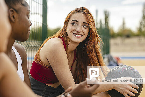 Smiling young woman holding basketball sitting with friends in basketball court