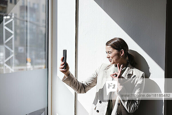 Businesswoman gesturing OK sign and taking selfie through mobile phone in office