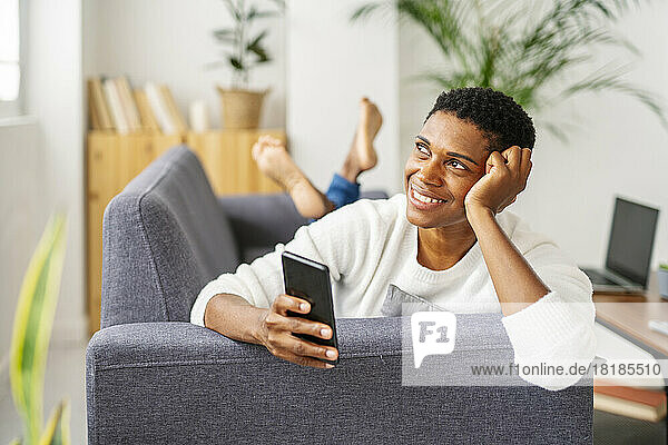 Smiling daydreaming woman lying on couch holding mobile phone