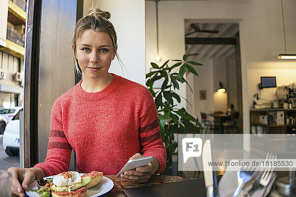 Smiling young freelancer sitting with mobile phone and plate of food at cafe