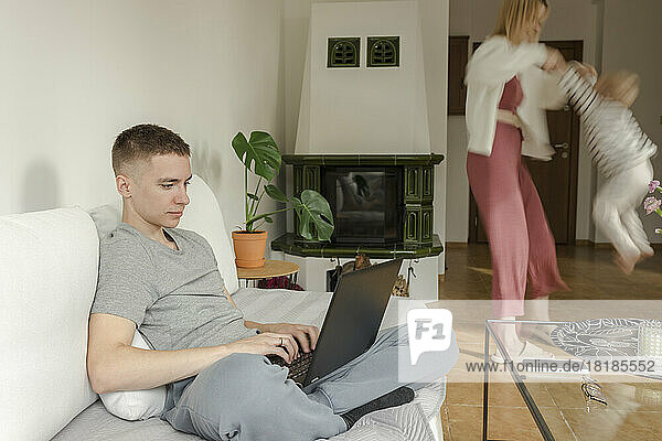 Father using laptop with mother and son playing in background