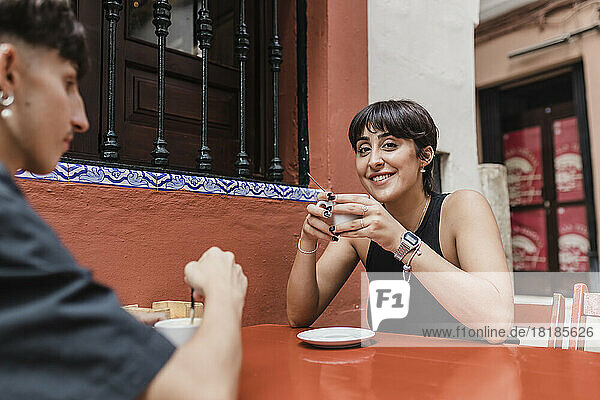 Lächelnde Frau hält Kaffeetasse in der Hand und sitzt mit ihrem Freund im Straßencafé
