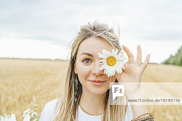 Lächelnde Frau bedeckt Auge mit weißer Blume im Feld