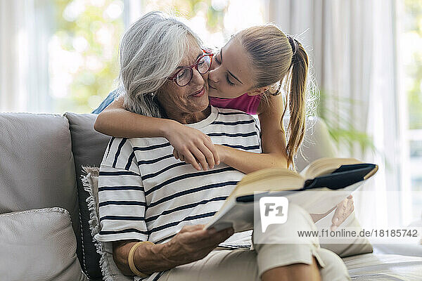 Girl kissing grandmother reading book sitting on sofa at home
