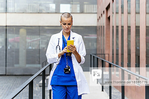 Mature doctor in lab coat using mobile phone amidst railings