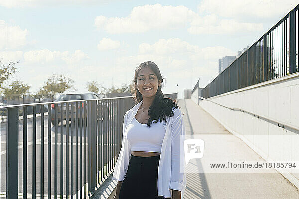 Smiling young woman standing at footpath by railing on sunny day