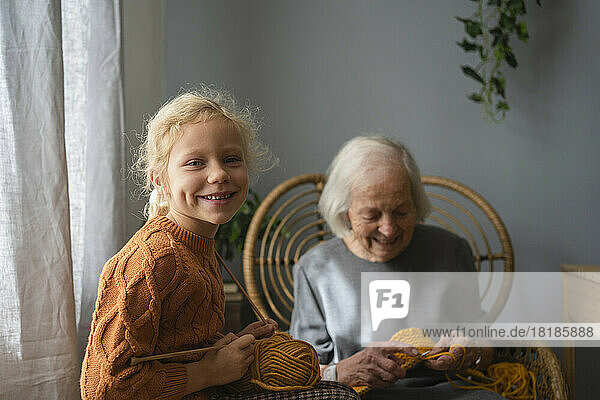 Happy girl holding ball of wool and knitting needle in front of grandmother at home