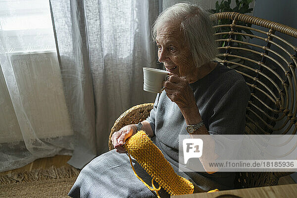 Senior woman having tea holding knitting wool on chair at home