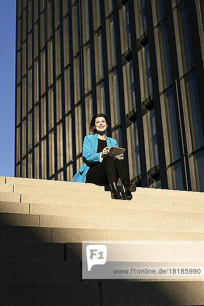Smiling businesswoman with tablet PC sitting near building on sunny day
