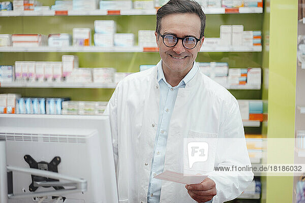 Happy pharmacist holding prescription in front of desktop PC at store