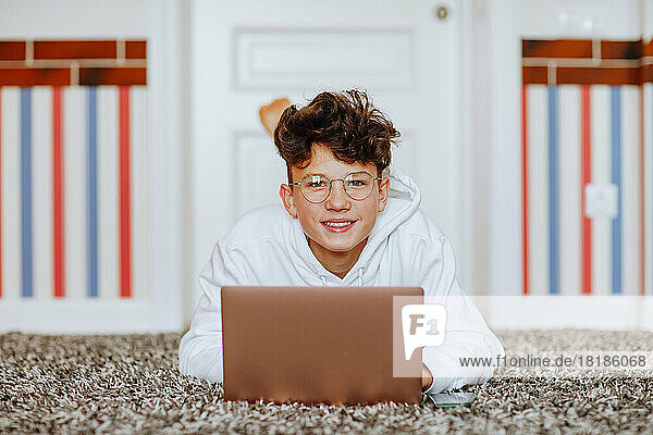 Smiling boy lying on carpet with laptop at home