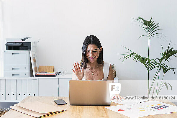 Smiling young businesswoman on video call waving at laptop in office