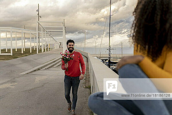 Young man walking with bouquet towards woman sitting on wall