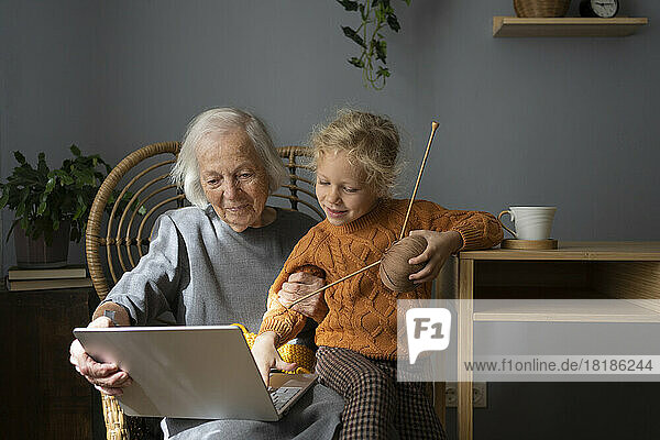 Smiling grandmother and granddaughter learning knitting through laptop at home