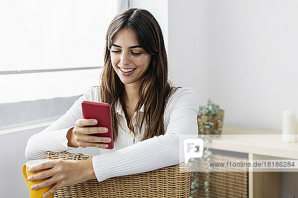Smiling woman holding coffee cup using smart phone on chair at home