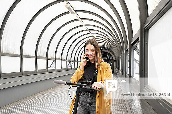 Smiling young woman talking on mobile phone standing with push scooter at footbridge