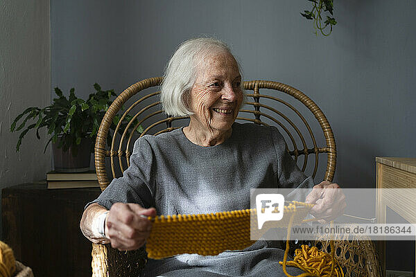 Happy senior woman with knitting wool and needle on chair at home