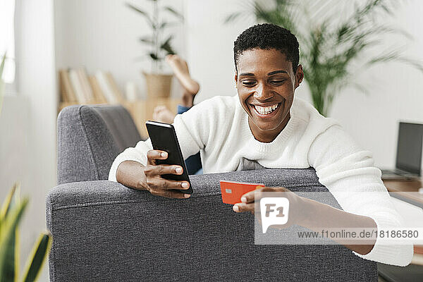 Lächelnde Frau liegt auf der Couch und benutzt Mobiltelefon und Kreditkarte