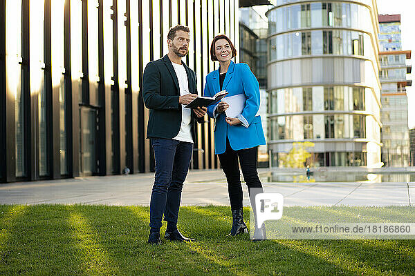 Businessman holding book discussing with colleague standing on grass in front of building