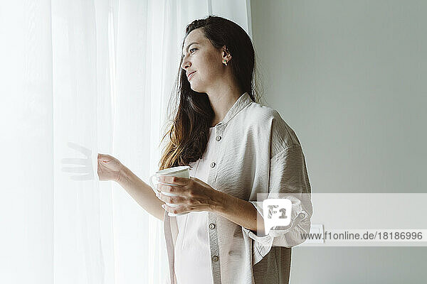 Nachdenkliche Frau mit Kaffeetasse und Blick durch das Fenster zu Hause
