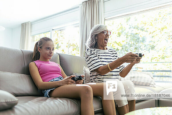 Excited grandmother playing video game with granddaughter sitting on sofa at home