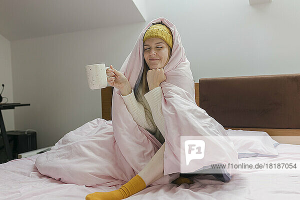 Young woman holding coffee cup wrapped in blanket on bed at home